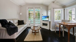 A living room with a sofa, coffee table, TV, and a dining area with a table and chairs. - Sage by Hyams Beach Seaside Cottages | Romantic, iconic heritage cottage (Hyams Beach)