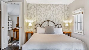 A bedroom with a large bed, two bedside tables, a wall-mounted lamp, and a window with shutters. - Sage by Hyams Beach Seaside Cottages | Romantic, iconic heritage cottage (Hyams Beach)