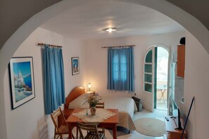 A bedroom with a bed, a desk, and a chair. There is a window with curtains, a door leading to another room, and a painting on the wall. - Akrotiri Flat near Red Beach (akrotiri)