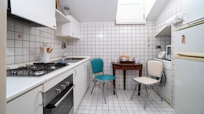 Cocina compacta con muebles blancos, estufa a gas, lavamanos y un pequeño comedor con dos sillas.