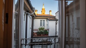 Blick durch ein Fenster auf ein Gebäude mit Ziegeldach, einen Balkon mit Blumentöpfen und einen Turm mit einer Spitze im Hintergrund.