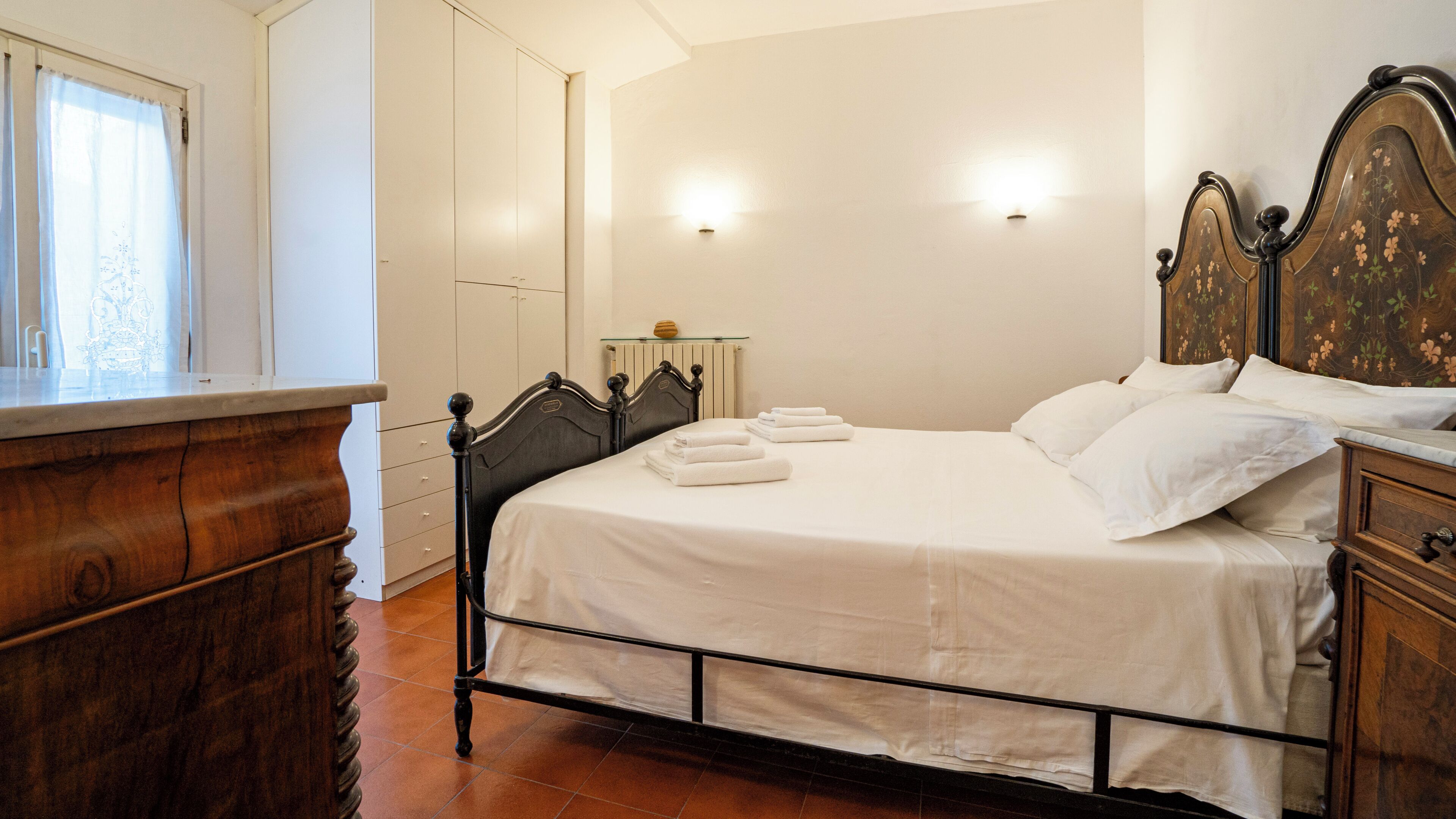 Una cama bien hecha con ropa de cama y toallas blancas, un aparador de madera y un radiador en una habitación con paredes blancas y una ventana.