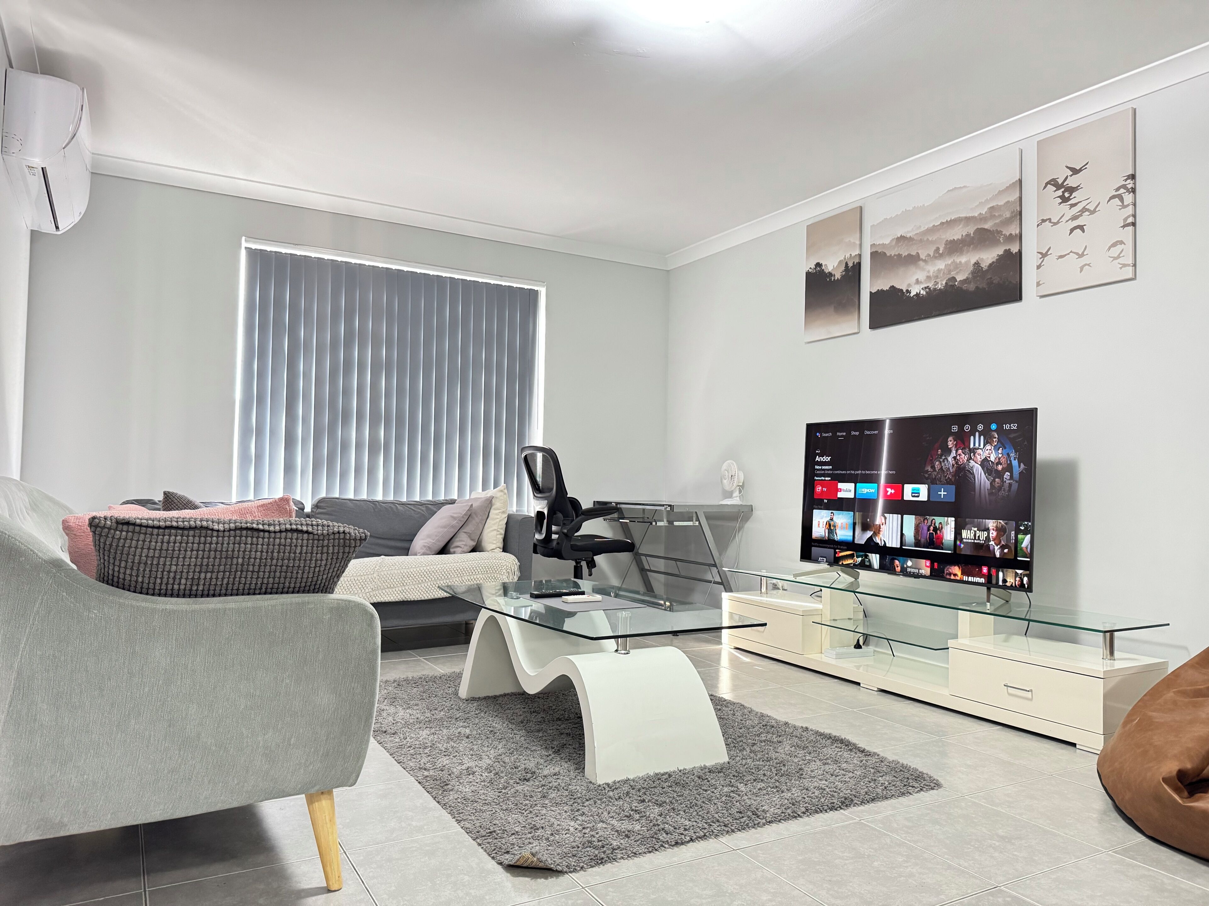 A modern living room with a sofa, armchair, glass coffee table, and a flat-screen TV displaying a streaming service interface.