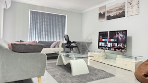 Una sala de estar moderna con un sofá, un sillón, una mesa de centro de vidrio y un televisor de pantalla plana mostrando la interfaz de un servicio de transmisión.