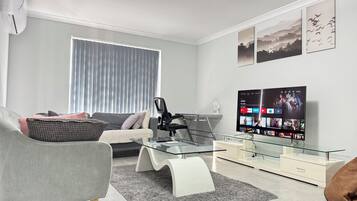 A modern living room with a sofa, armchair, glass coffee table, and a flat-screen TV displaying a streaming service interface.