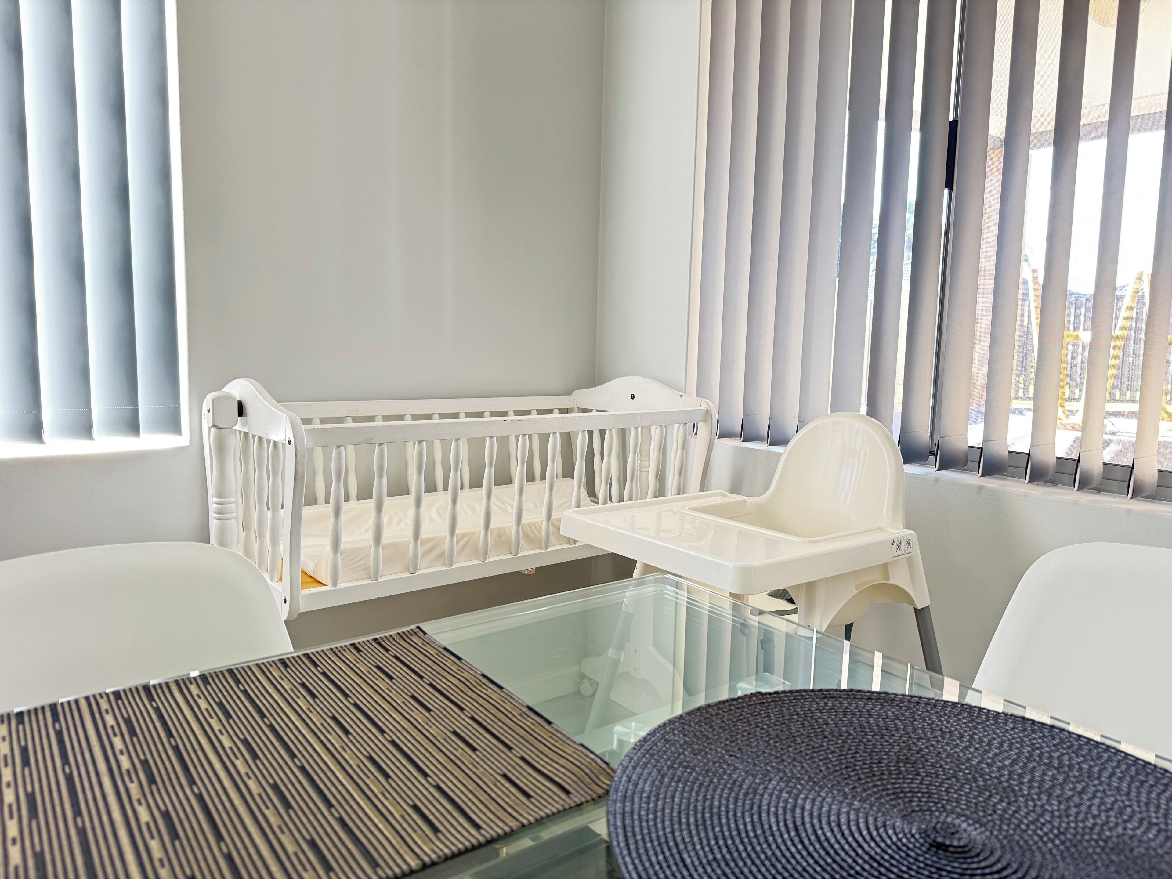 A baby crib with a changing table beside it, a glass table with a woven placemat, and a view through a window with blinds.