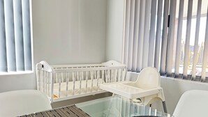 Una cuna para bebé con un cambiador a su lado, una mesa de vidrio con un mantel tejido, y vistas a través de una ventana con persianas.