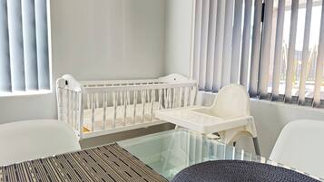 A baby crib with a changing table beside it, a glass table with a woven placemat, and a view through a window with blinds.