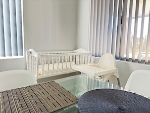 A baby crib with a changing table beside it, a glass table with a woven placemat, and a view through a window with blinds. - Blissful Haven (Perth)