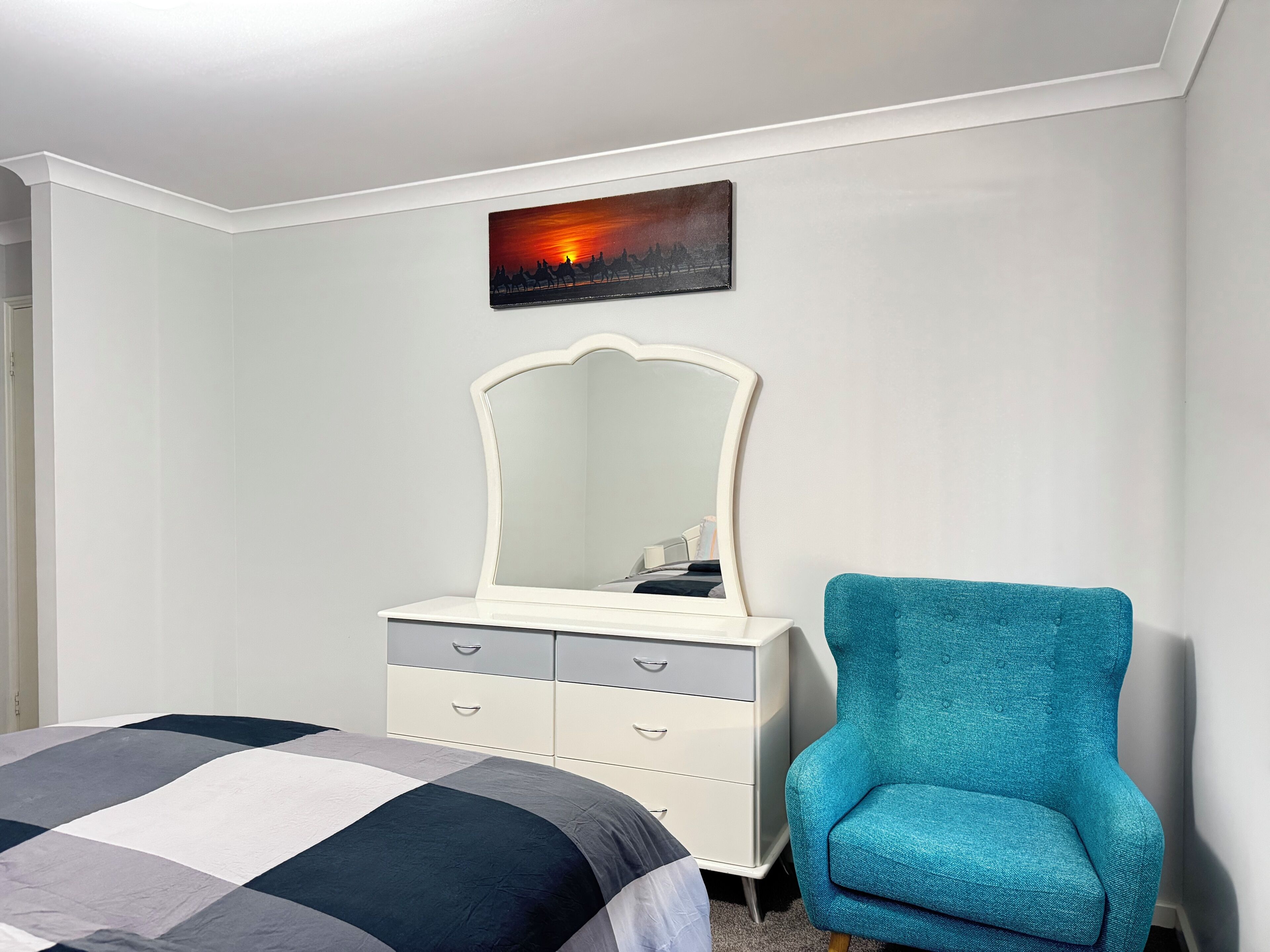 A bedroom with a bed, a dresser with drawers, a chair, a wall-mounted mirror, and a painting of a sunset.