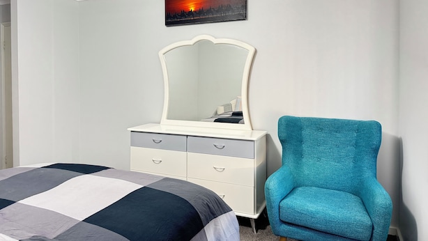 Un dormitorio con cama, cómoda con cajones, silla, espejo en la pared y una pintura de un atardecer.
