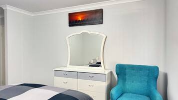 A bedroom with a bed, a dresser with drawers, a chair, a wall-mounted mirror, and a painting of a sunset.