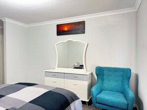 Une chambre à coucher comprenant un lit, une commode avec des tiroirs, une chaise, un miroir fixé au mur et un tableau représentant un coucher de soleil.