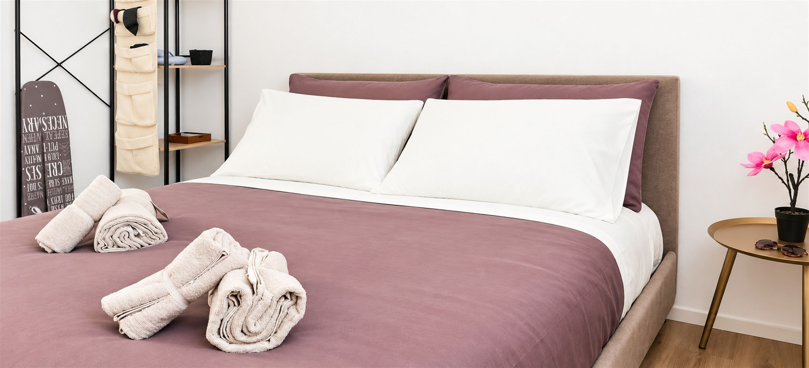 Een netjes opgemaakt bed met wit en roze beddengoed, twee opgerolde handdoeken, een klein rond tafeltje met zonnebril en een potplant met roze bloemen.