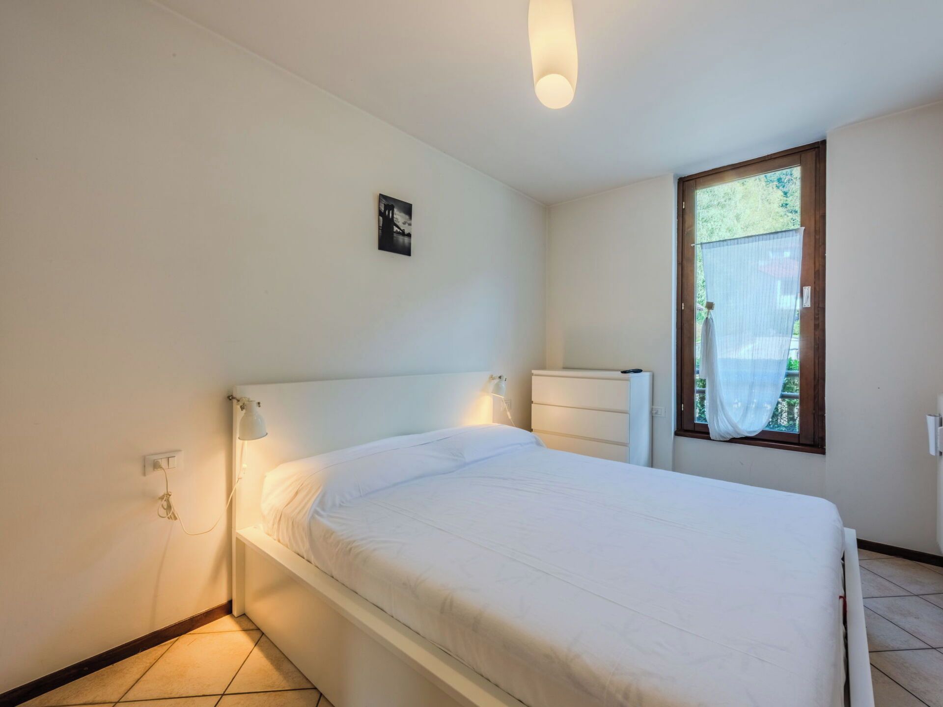 Een eenpersoonskamer met een wit hoofdeinde, een nachtkastje, een wandlamp en een raam met gordijn.