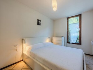 Ein Einzelzimmer mit einem weißen Kopfende, einem Nachttisch, einer Wandlampe und einem Fenster mit Vorhang.