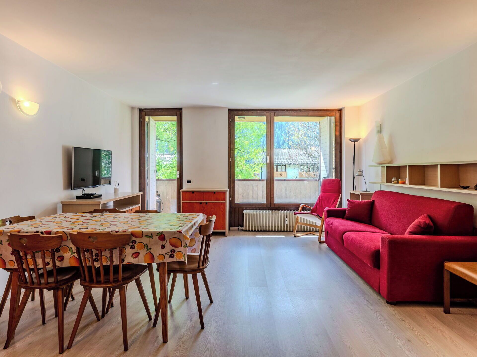 Een woonkamer met een rode bank, een eettafel met stoelen, een televisie en grote ramen.