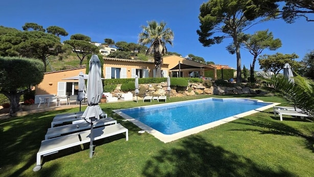 Una piscina amb gandules i ombrelles, envoltada per gespes i arbres ben cuidats, i un edifici al fons.