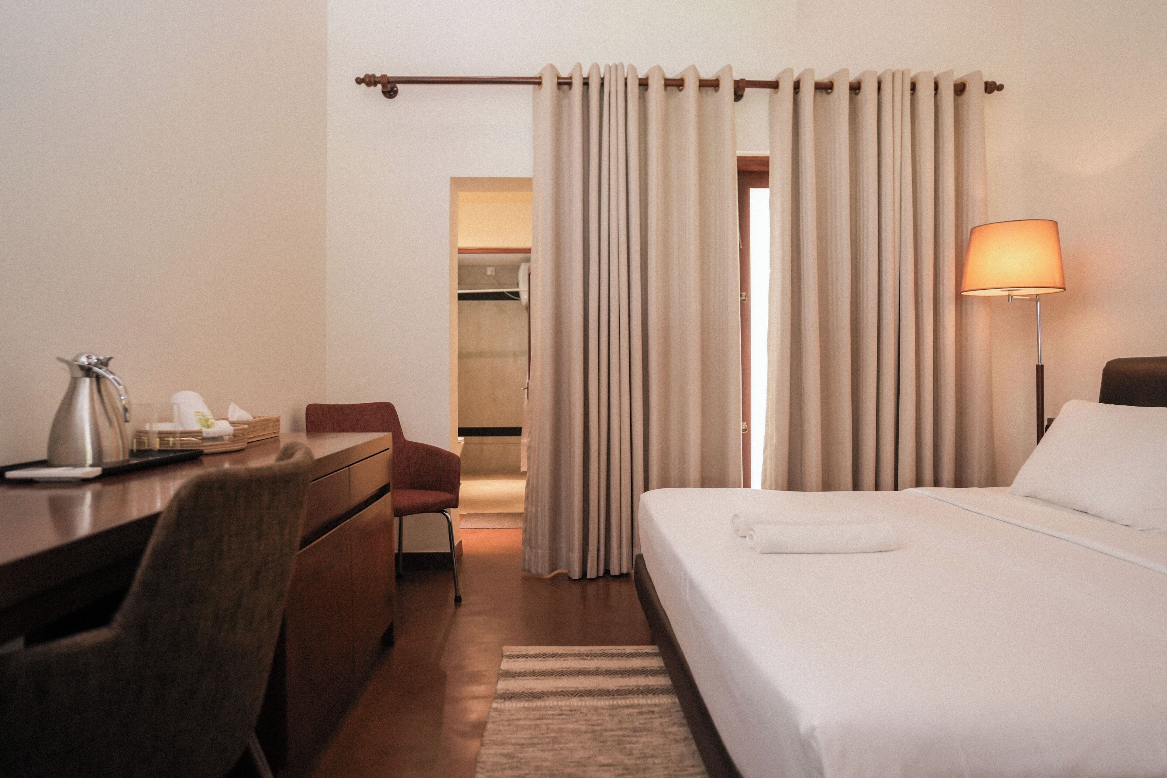 Habitación de hotel con cama, escritorio, silla, lámpara y baño visible a través de una cortina.