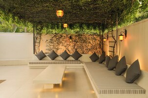 Un espacio exterior moderno con asientos, un sofá blanco y cojines, un banco e iluminación instalada en la pared.