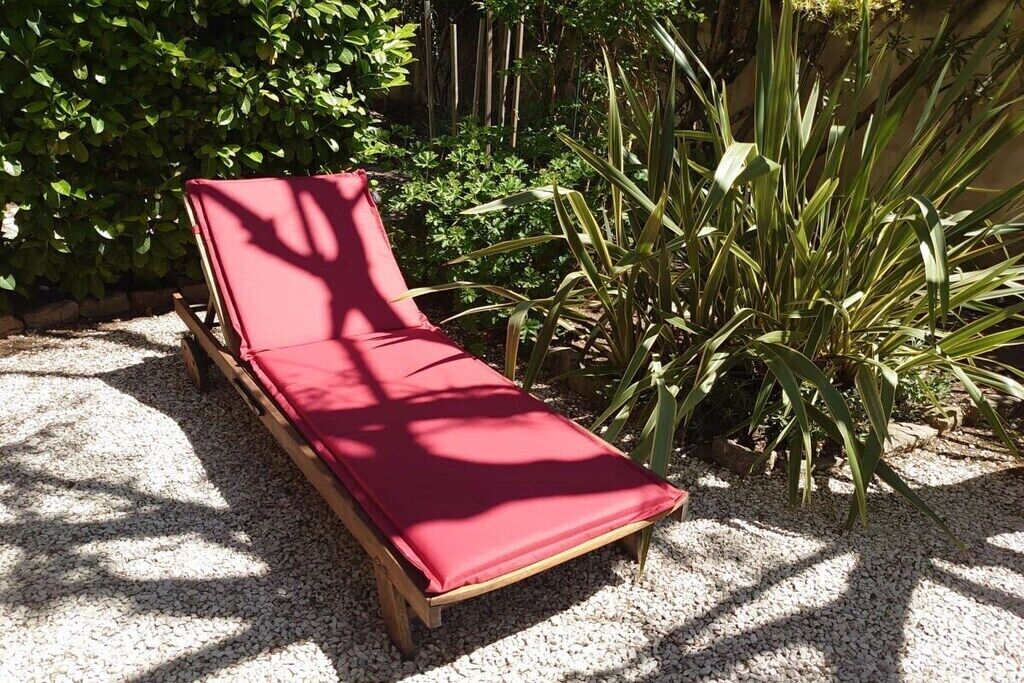 A wooden lounge chair with a red cushion, positioned on a gravel surface, surrounded by greenery.