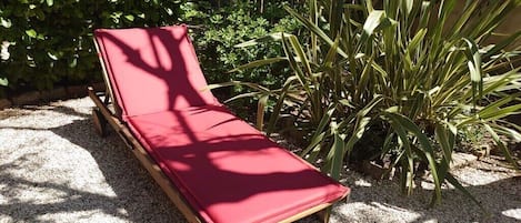 A wooden lounge chair with a red cushion, positioned on a gravel surface, surrounded by greenery.