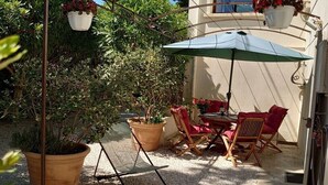 Pátio externo com mesa e cadeiras, rodeado por plantas em vasos e um cachepot pendente.