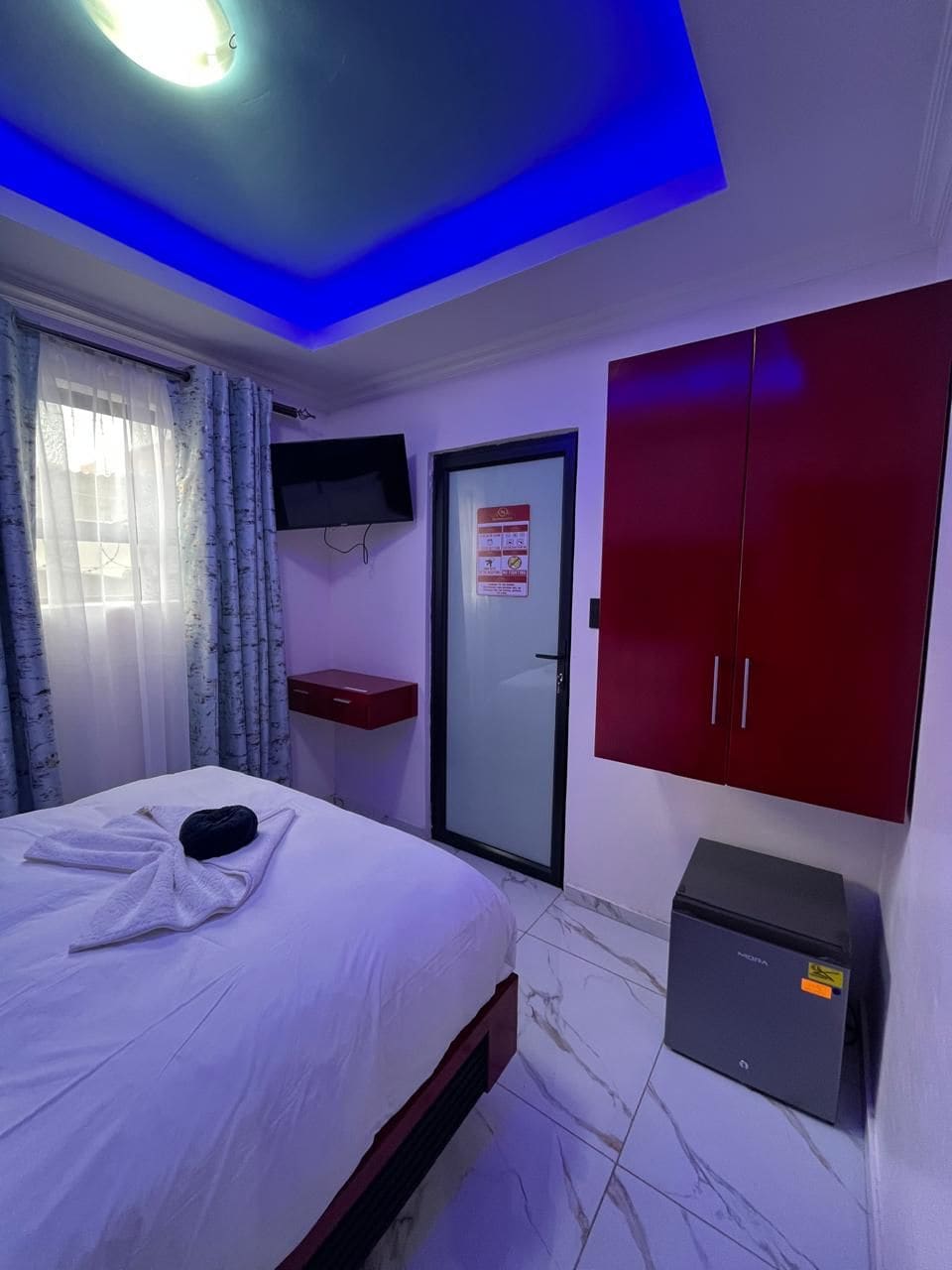 Una habitación de hotel con una cama, un televisor montado en la pared, un armario rojo y un mueble pequeño.