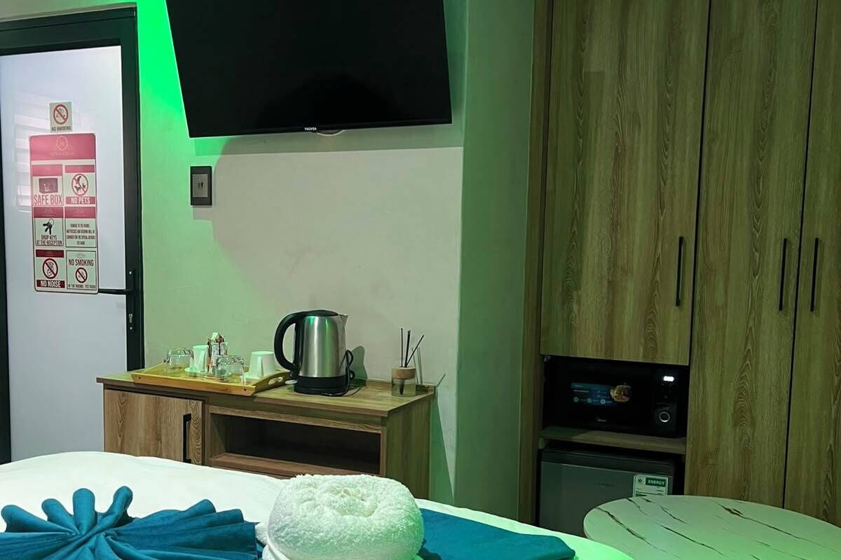 A hotel room with a bed, a flat-screen TV mounted on the wall, a wooden wardrobe, a small table with a kettle, and a safe box on the wall.