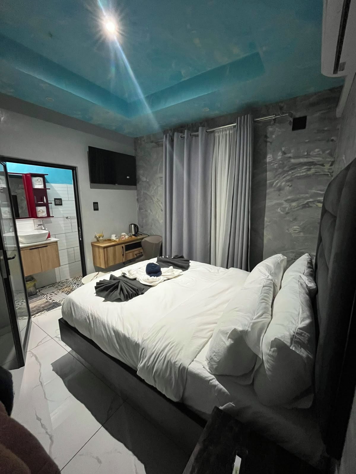 A modern bedroom with a bed, a bedside table, a mirror, and a bathroom visible through a glass door.