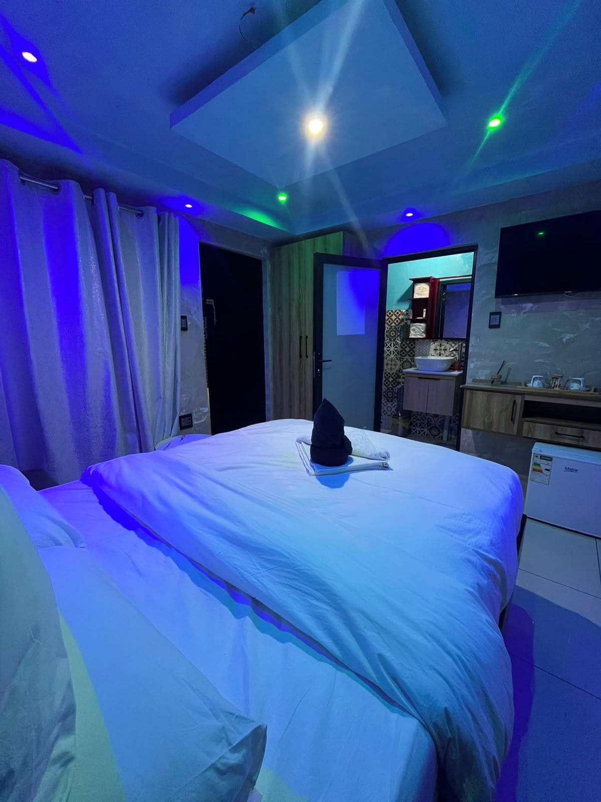 A hotel room with a bed, a flat-screen TV mounted on the wall, and a small kitchenette area.