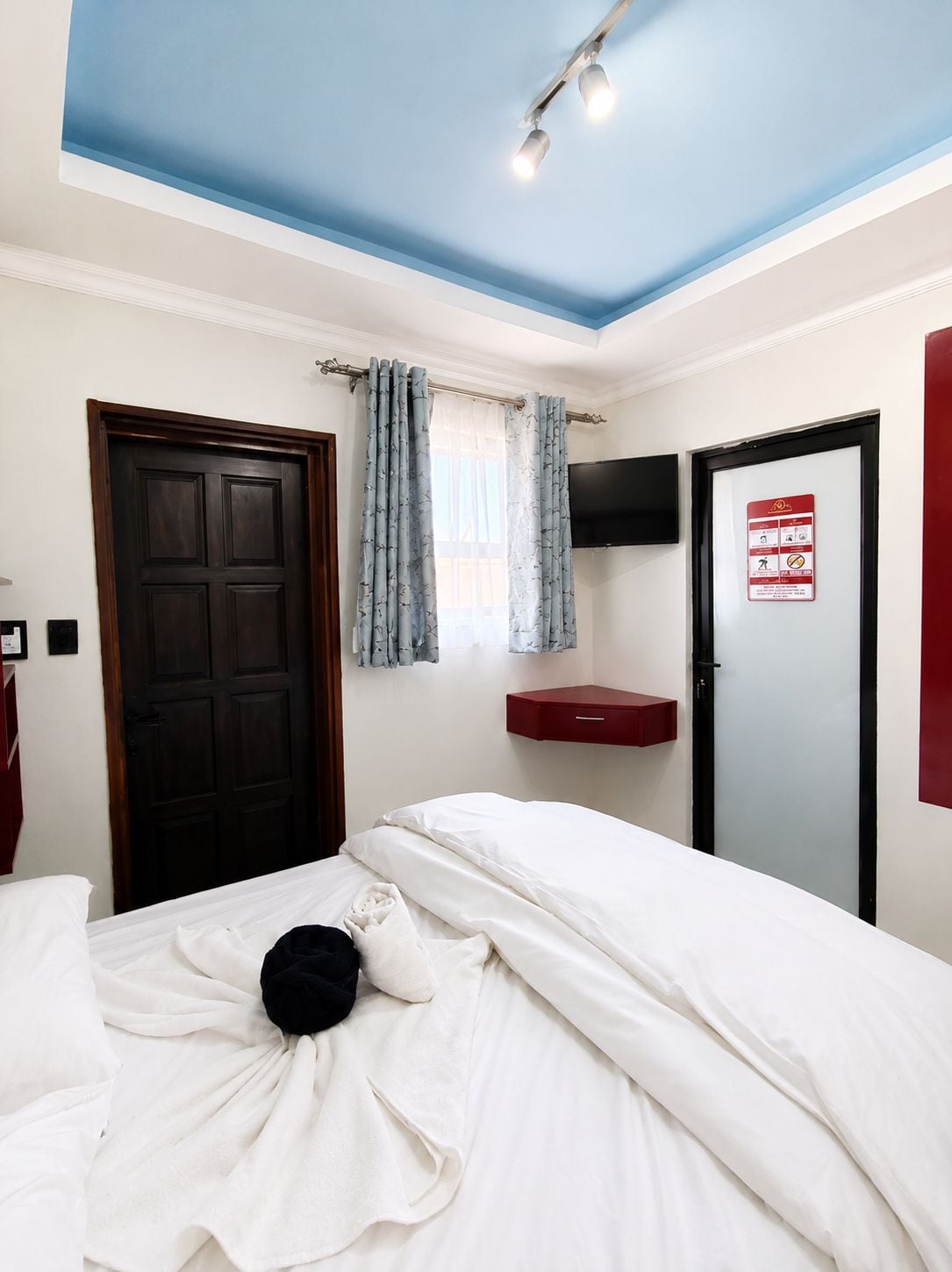 Un dormitorio con cama, una puerta, una ventana con cortinas, un televisor montado y un armario rojo.