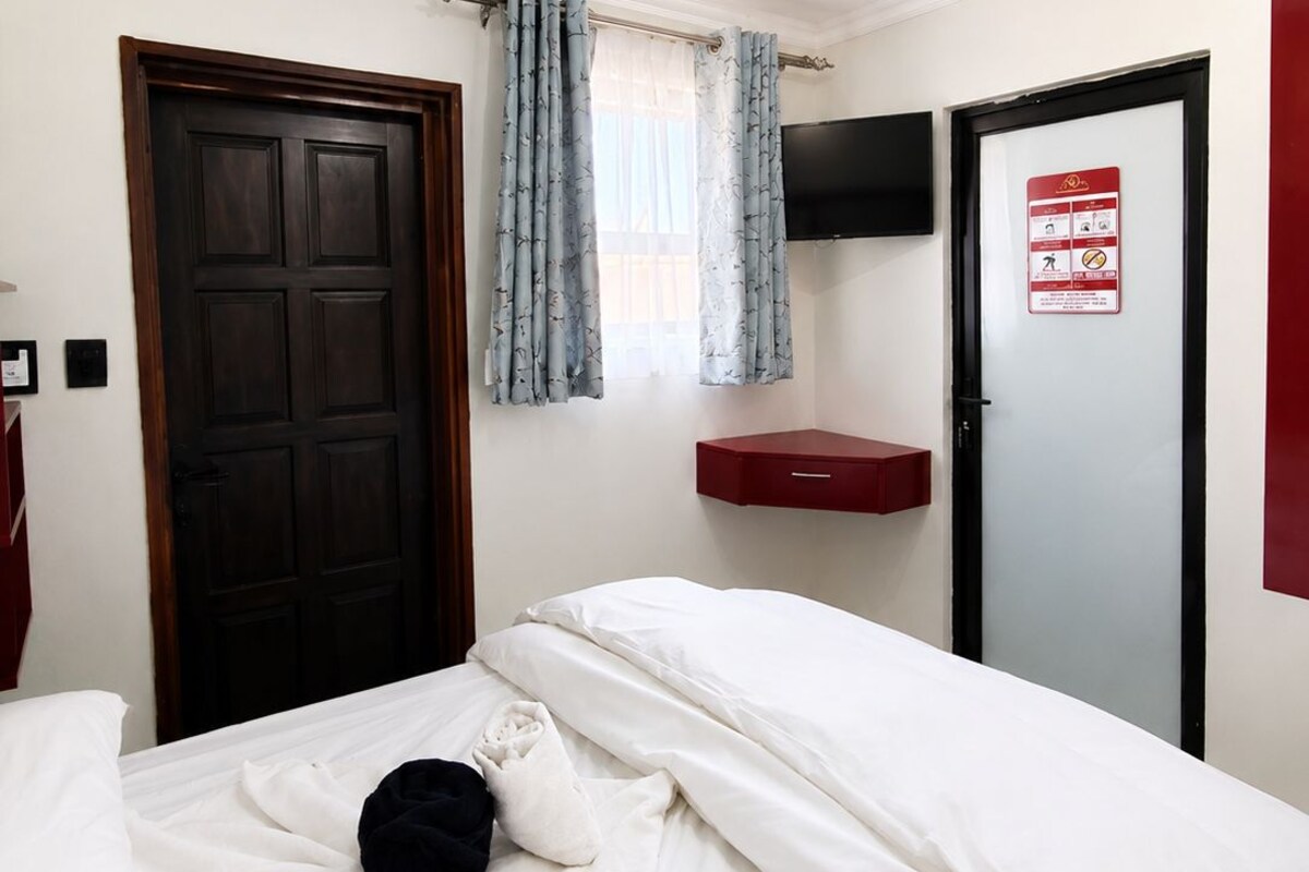 A bedroom with a bed, a door, a window with curtains, a mounted television, and a red cabinet.