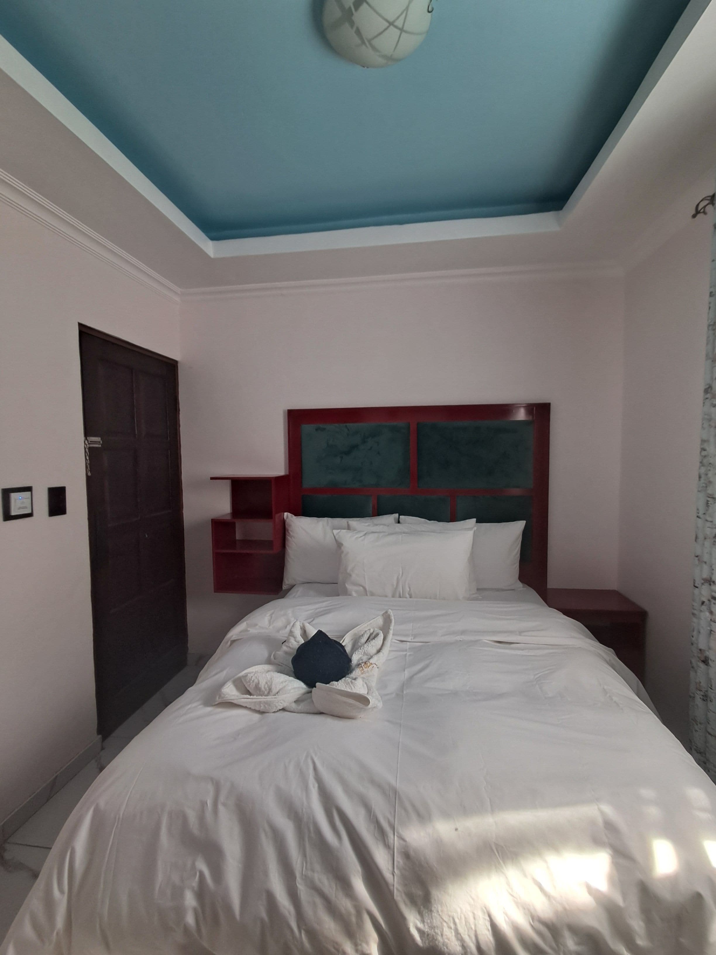 A hotel room with a bed, a nightstand, a red headboard, and a door.