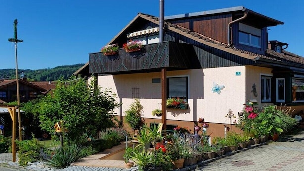 Une maison à deux étages, dotée d’un toit en pente, d’un balcon orné de jardinières et d’un jardin où poussent diverses plantes et fleurs.