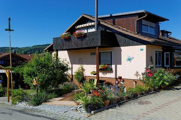 Een tweelaags huis met een schuin dak, een balkon met bloembakken, en een tuin met diverse planten en bloemen.