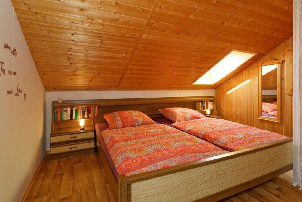 Un dormitorio con techo inclinado, paredes de madera, una cama con ropa de cama estampada, una mesita de noche y una estantería con libros.