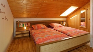 Une chambre avec un plafond en pente, des murs en bois, un lit avec une literie à motifs, une table de chevet et une étagère remplie de livres.