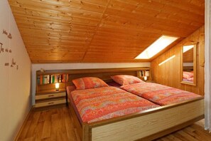 Een slaapkamer met een schuine kap, houten wanden, een bed met beddengoed met een patroon, een nachtkastje en een plank met boeken.