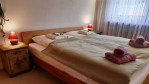 Una cama individual con cabecera de madera, dos lámparas de noche, una mesita de noche con cajón y una ventana con cortinas traslúcidas.