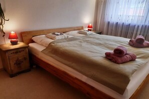 Una cama individual con cabecera de madera, dos lámparas de noche, una mesita de noche con cajón y una ventana con cortinas traslúcidas.