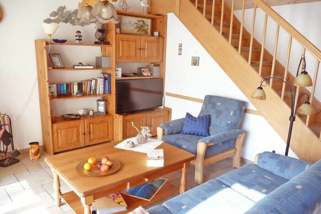 Uma sala de estar com escada de madeira, uma poltrona azul, uma mesa de centro de madeira com frutas e uma estante de livros.