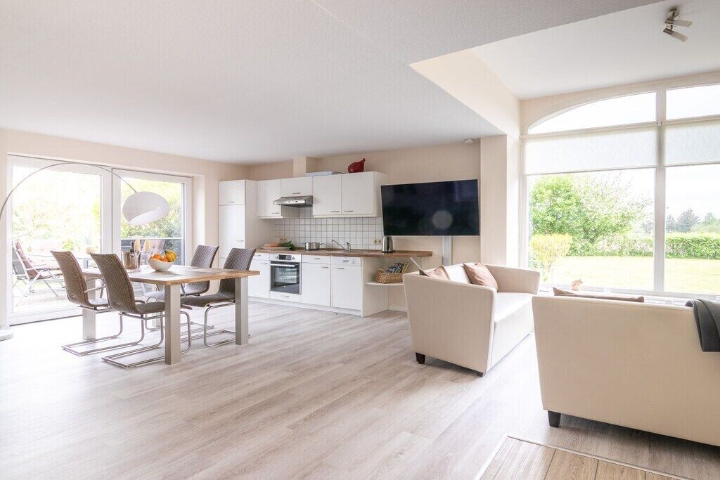 Een moderne keuken met witte kasten, een eethoek met tafel en stoelen, en een groot raam met uitzicht op groen.