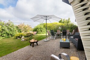 Een terras buiten met een tafel, stoelen, een grote parasol en een vuurkuil.