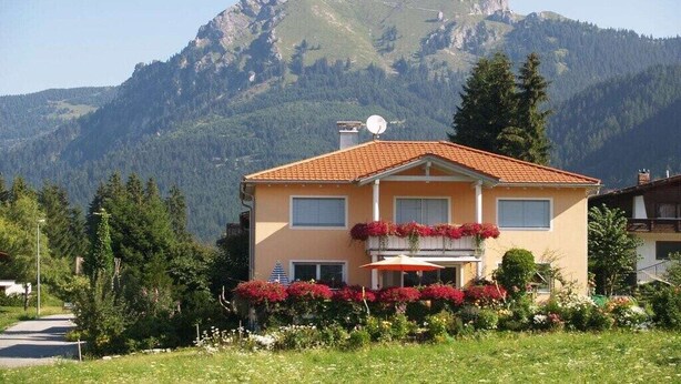A two-story building with a red-tiled roof, a balcony with red flowers, and a mountain backdrop. - Holiday apartment in Grän with swimming pool (Grän)