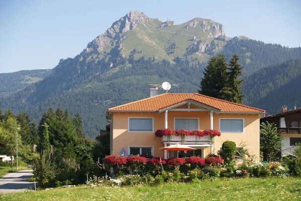 A two-story building with a red-tiled roof, a balcony with red flowers, and a mountain backdrop. - Holiday apartment in Grän with swimming pool (Grän)