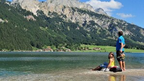 A lake with mountains in the background, a person standing, and another person sitting on the shore. - Holiday apartment in Grän with swimming pool (Grän)