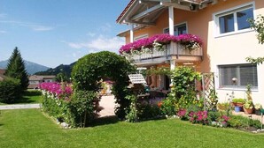A two-story house with a balcony adorned with flowers, a well-maintained garden, and a mountain in the background. - Holiday apartment in Grän with swimming pool (Grän)