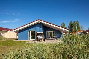 Een blauw huis met een rood dak, een terras met een tafel en stoelen, en een gazon ervoor.