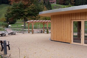 A building with a wooden exterior and a glass door, situated in a park with benches and a pergola. - Holiday apartment with 2 bedrooms (Oberstaufen)